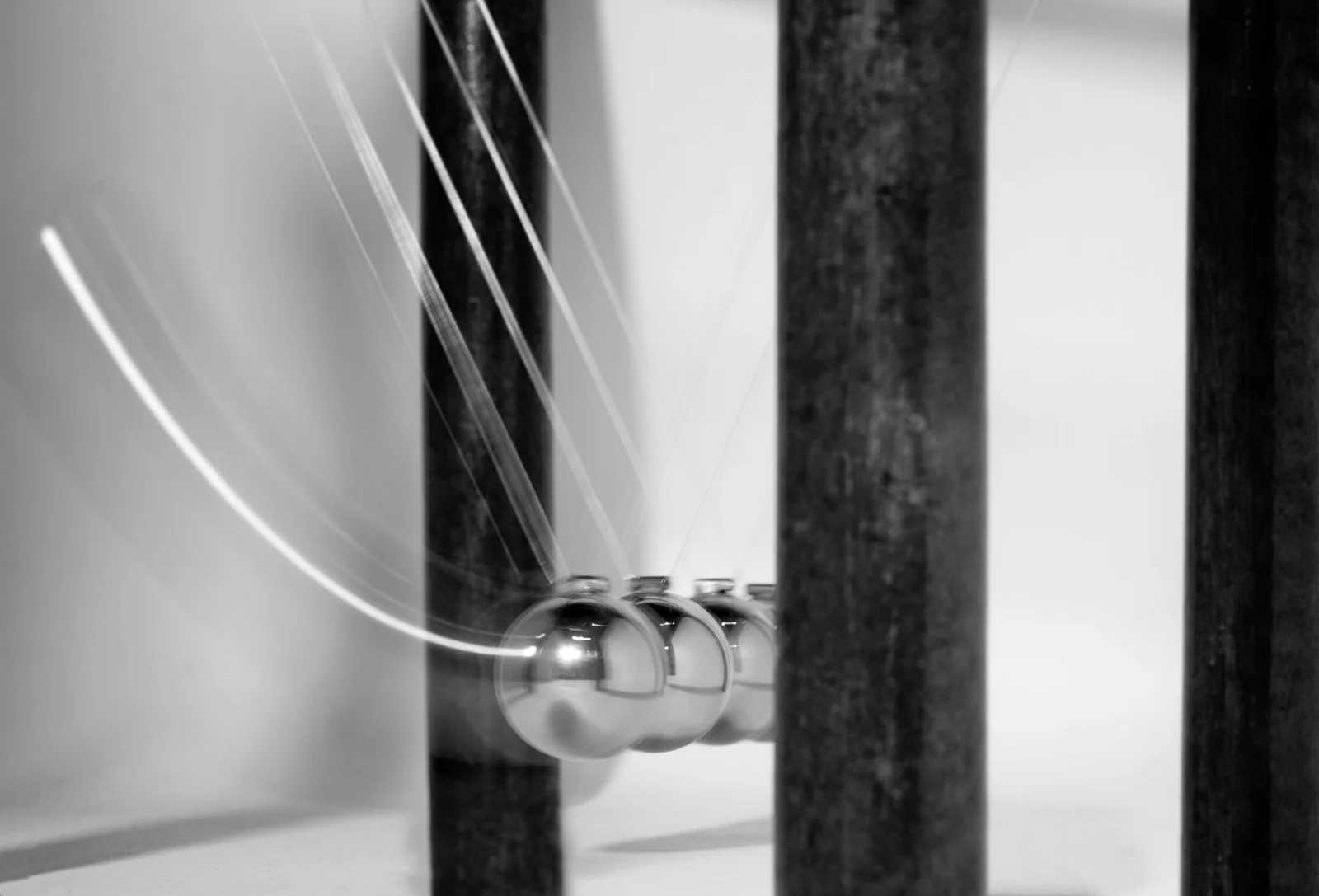 검은 기둥 사이에 매달린 금속 구 여러 개와 왼쪽 끝 구가 왕복한 흰 곡선 궤적이 보이는 흑백 장노출 사진